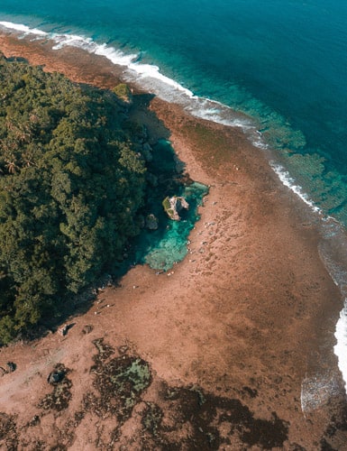 aerial view of the coastline on Siargao Island, Philippines. Golden brown sand covers a crescent shaped part of the land, where turquoise water meets the shore.