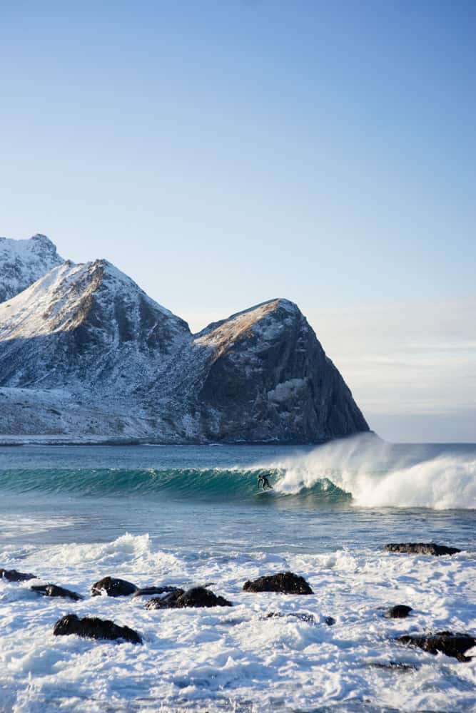 Photographie de surf en Norvège ; un surfeur vêtu d'une combinaison de plongée surfe dans l'eau froide au pied d'une montagne.