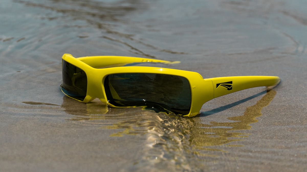 Die schwimmende Sonnenbrille 'Flo' von LiP in Senfgelb am Strand mit darüber fließendem Wasser