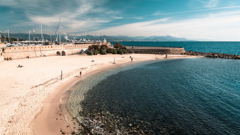 Der Strand von Antibes mit seinem sichelförmigen Strand und dem blauen Wasser, das an seine Ufer stößt. Im Hintergrund dümpeln die Yachten des Port Vauban im Wasser, und dahinter sind die schneebedeckten Berge zu sehen