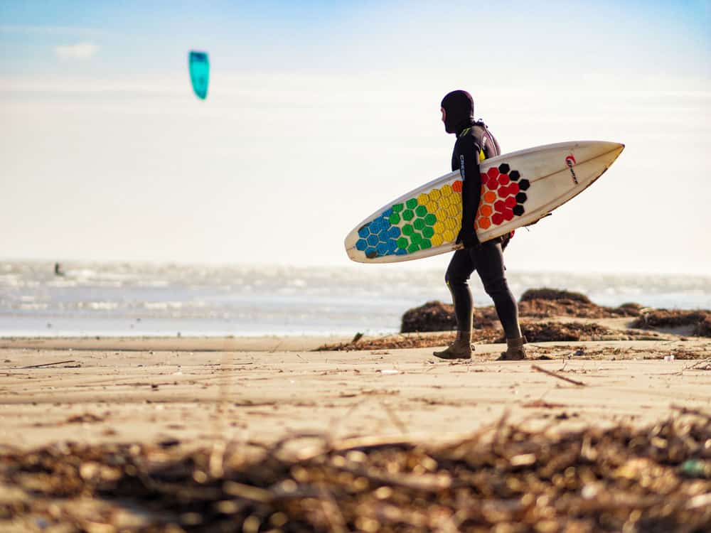 Mann mit Vollkapuzen-Neoprenanzug trägt Surfbrett am Strand beim Kitesurfen