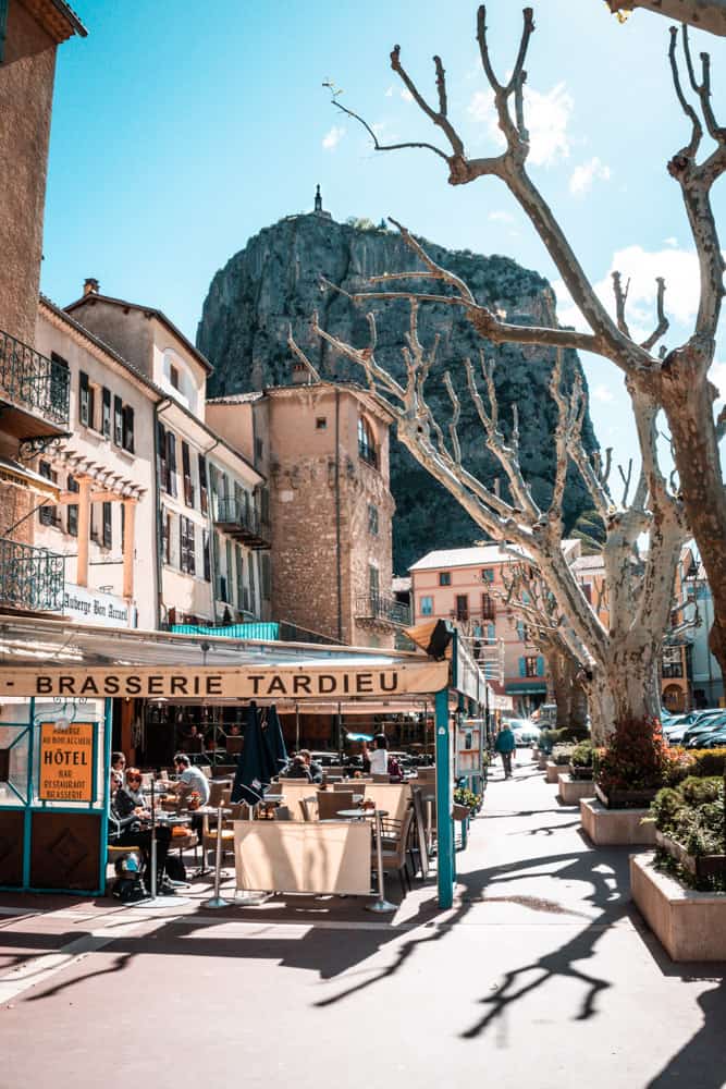 Die Stadt Castellane auf Straßenniveau, Südfrankreich. Im Vordergrund befinden sich kalkhaltige Buchen und ein Café (Brasserie). Im Hintergrund sieht man eine hohe Klippe mit einer Kapelle auf dem Gipfel.