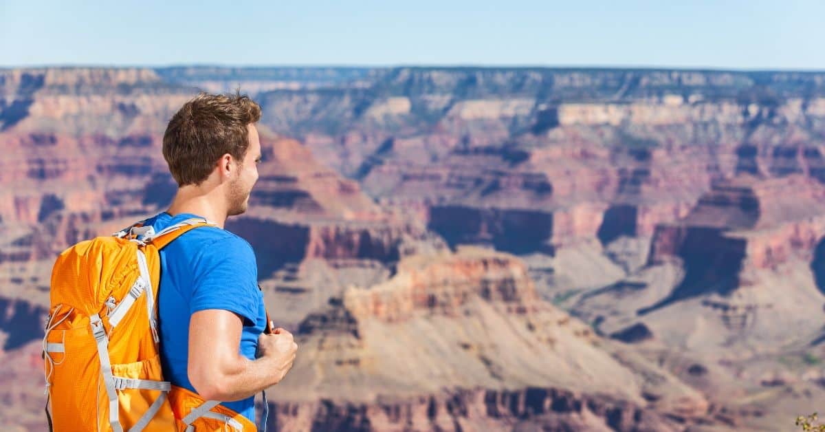 sehr scharfes Bild eines jungen Mannes, der ein leuchtend blaues T-Shirt und einen orangefarbenen Rucksack trägt, als er zu einer Tageswanderung durch den Grand Canyon von Rim zu Rim aufbricht. Hinter ihm sehen wir die leuchtend blaue und rote Landschaft des Grand Canyon.