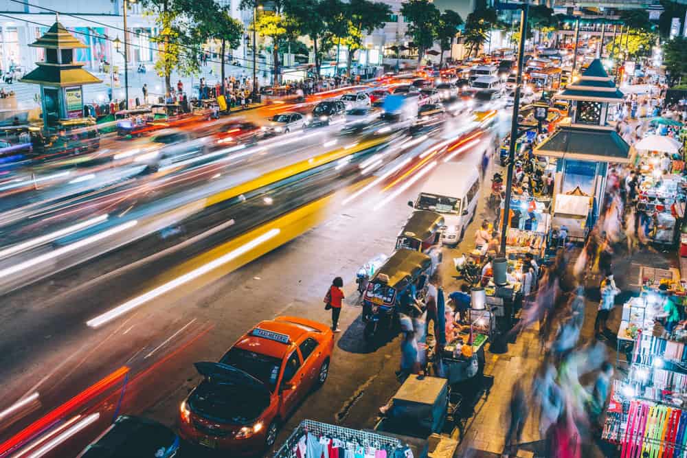 Long exposure at nighttime on a busy street in Bangkok. we see the blur of fast moving cars driving down the street and a bustling night market on the right.