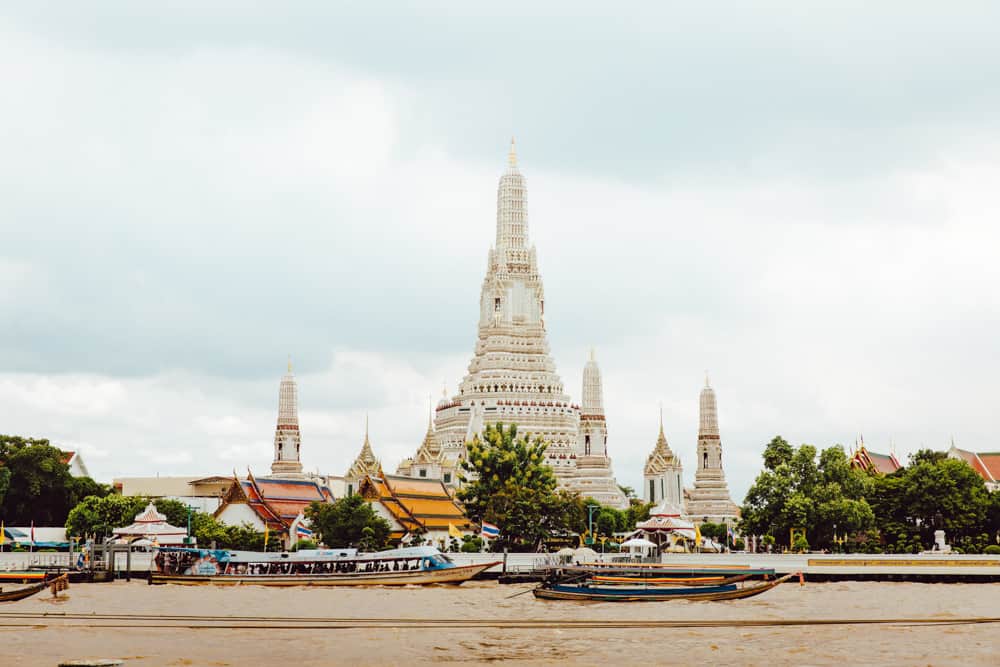 Een enorme witte tempel in Bangkok, met op de voorgrond de rivier vol kleurrijke sloepen. Bewolkte grijze lucht erboven.