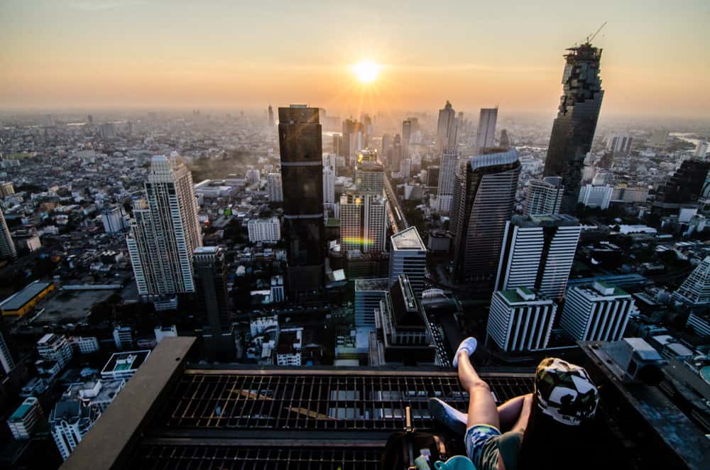 Sonnenaufgang über Bangkok: eine Frau im Vordergrund sitzt auf einem Dach und blickt auf die Wolkenkratzer der Stadt