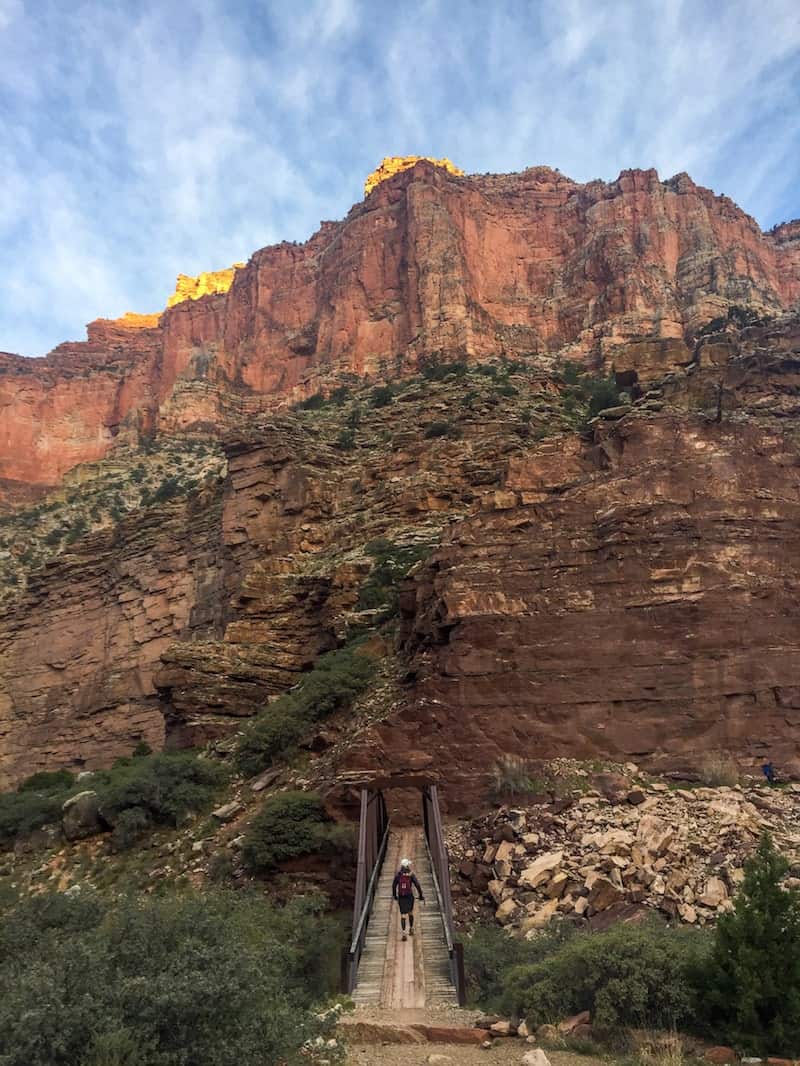 blue sky creeping over the top of the red rocky cliffs of the grand canyon, with a very small hiker just visible in the foreground