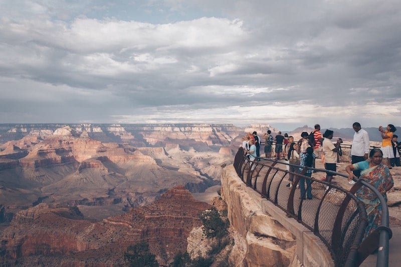 a gaggle of people stand on the right side of the frame on top of a viewpoint area, on the left side we see the hazy landscape of the grand canyon sprawling in every direction