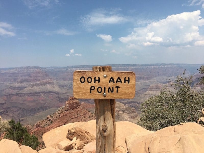 een wegwijzer op de voorgrond met de tekst "ooh aah point" en op de achtergrond het wazige landschap van de grand canyon op een zonnige dag