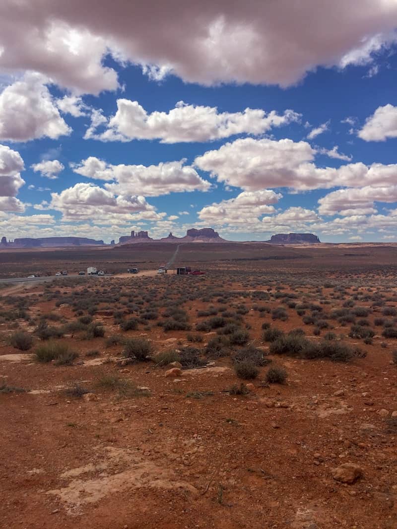 top of the picture is filled with blue sky and cotton candy clouds. on the horizon, the peaks of the grand canyon lay in wait. the bottom half of the picture is just red dirt and some rocks, with scraggly green tufts of grass growing here and there.