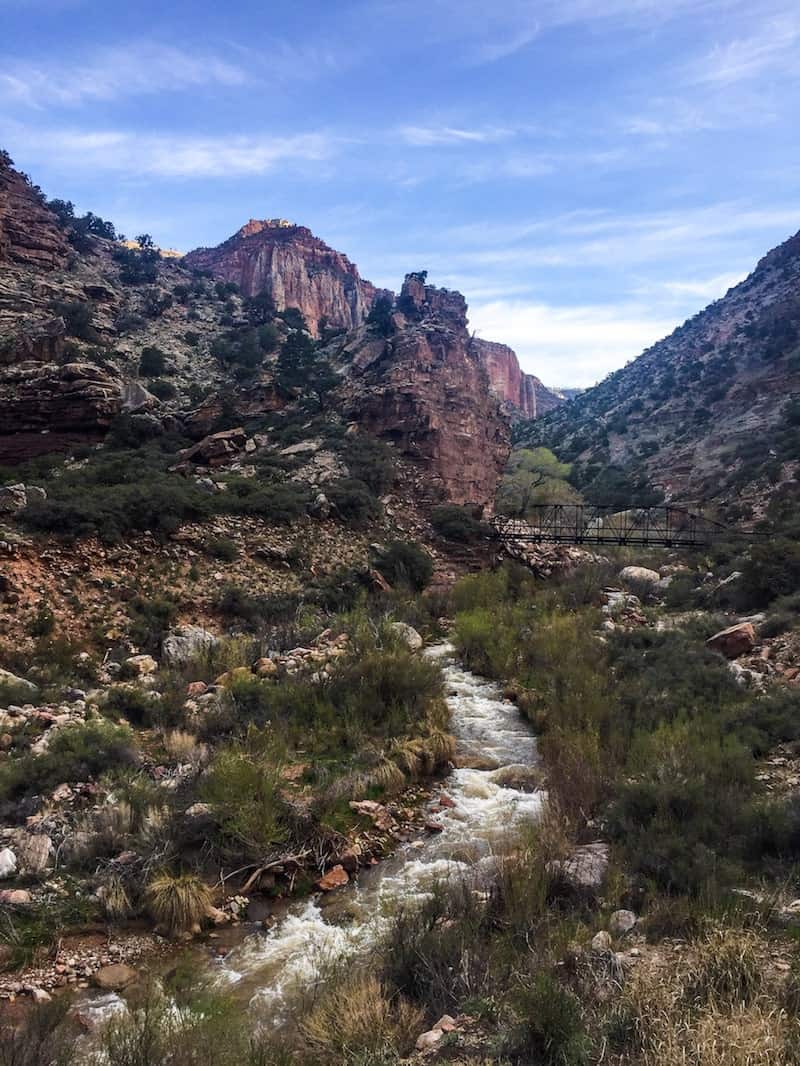 Un ciel bleu plane sur le paysage du Grand Canyon ; le soleil semble être derrière un nuage et les couleurs sont un peu atténuées. Une rivière coule au centre de l'image, au premier plan.