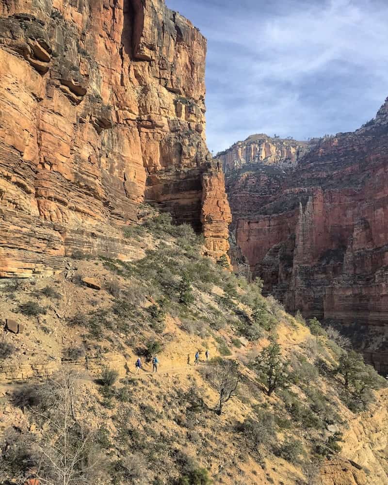 vertical image of the super high rock faces of the grand canyon. there is a very small path leading from left to right, with 6 people hiking along the ridge.