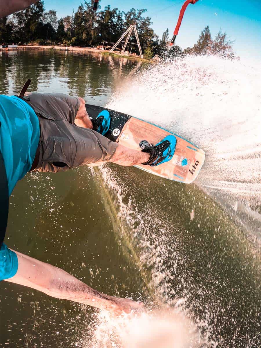 Vertikales Bild eines Mannes beim Wakeboarden in einem Cable Park. Das Bild zeigt den Unterleib und die Beine des Fahrers, der ein hellblaues T-Shirt, khakifarbene Shorts und passende blaue Wakeboots trägt. Seine Hand berührt das spritzende Wasser im See.