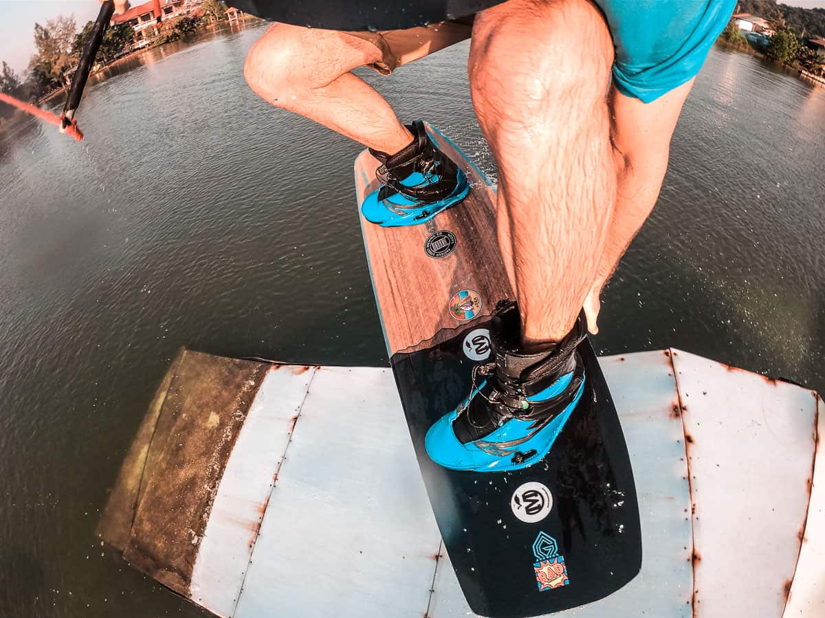 Jims Beine, während er Cable Wakeboarding betreibt. Er trägt blaue Ronix-Stiefel und fährt ein hölzernes und schwarzes Liquid Force Board. Er kommt von der Spitze eines Kickers und greift sein Brett