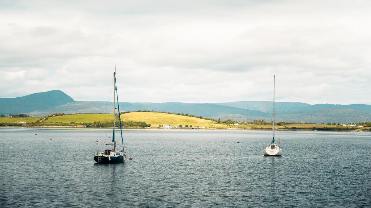 whiddy island on an overcast day, with two sailboats floating in the bay in the foreground