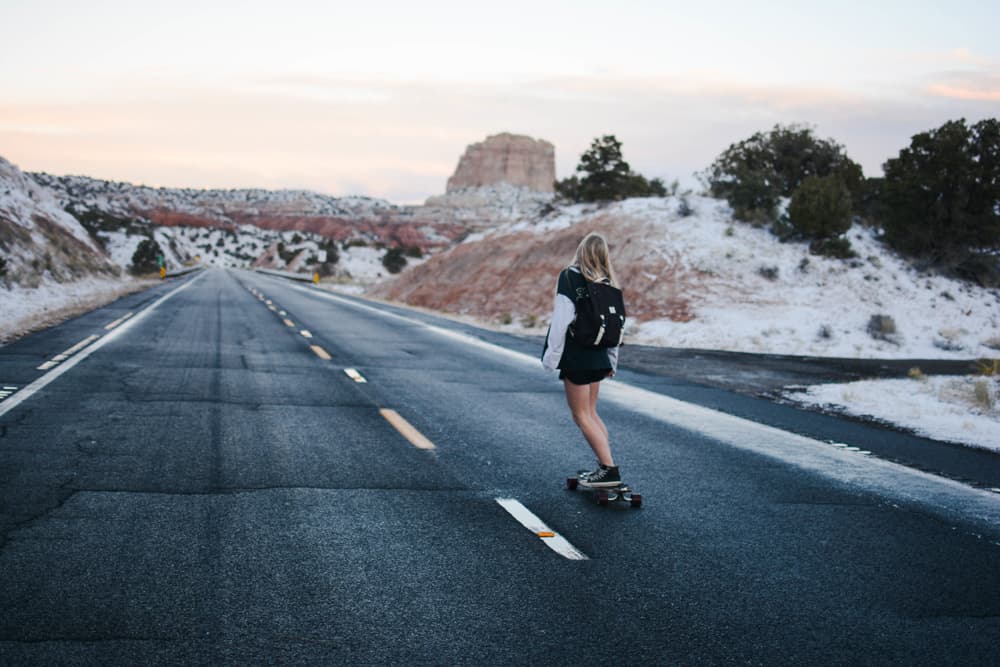 skate feminino descendo estrada asfáltica com afloramentos rochosos lavados no fundo
