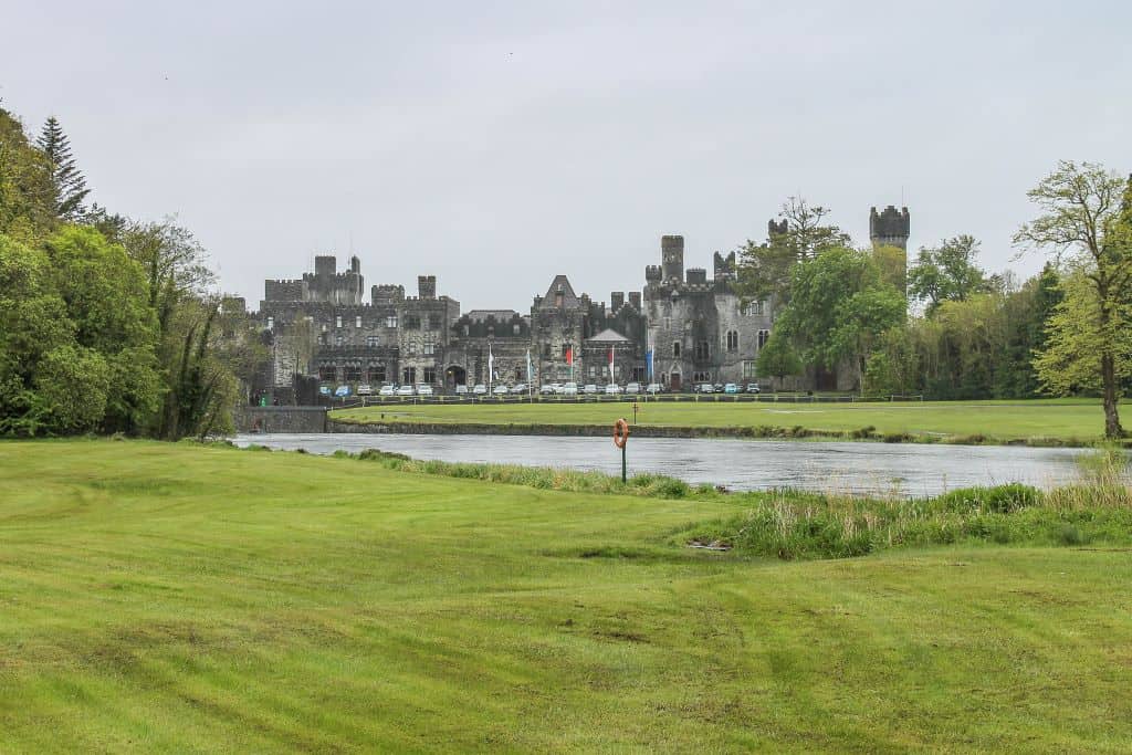 grassy manicured lawn in the foreground with ashford castle sitting just beyond a silvery pond in the background. a grey sky hangs overhead.