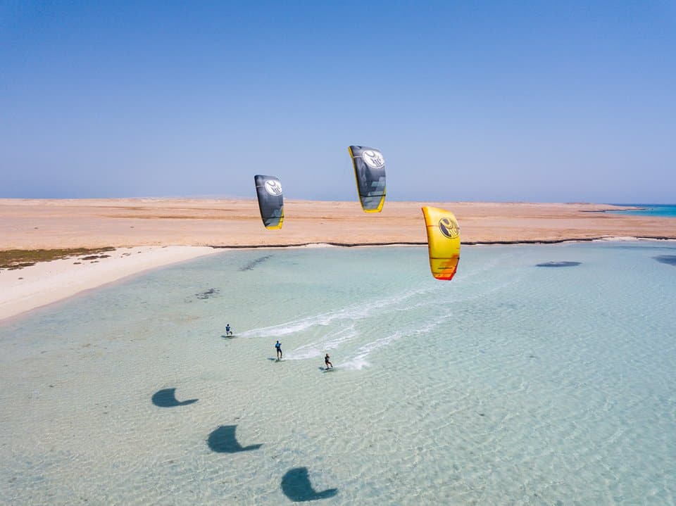 trois kitesurfers surfent à l'unisson sur les eaux azures d'el gouna, en égypte, avec le désert en arrière-plan et un ciel bleu clair au-dessus.