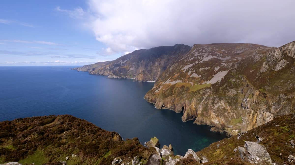 ruhiges blaues wasser links trifft auf die rostigen und grasfarbenen berge von slieve league. zwischen den klippen im vordergrund erscheinen die schwachen farben eines regenbogens.