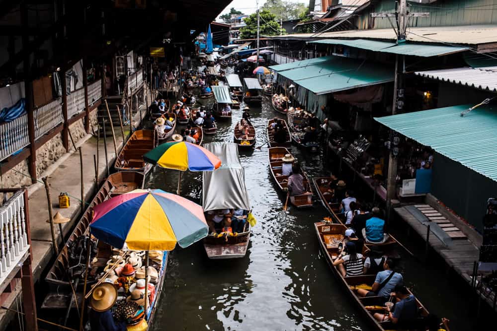 een van de drijvende markten van bangkok van bovenaf gezien. het kanaal is druk bevaren met kleine bootjes -- sommige met toeristen aan boord en andere met verkopers en hun waren. groene daken bedekken een deel van het kanaal rechts, terwijl regenboogkleurige paraplu's sommige boten links beschermen tegen het felle zonlicht