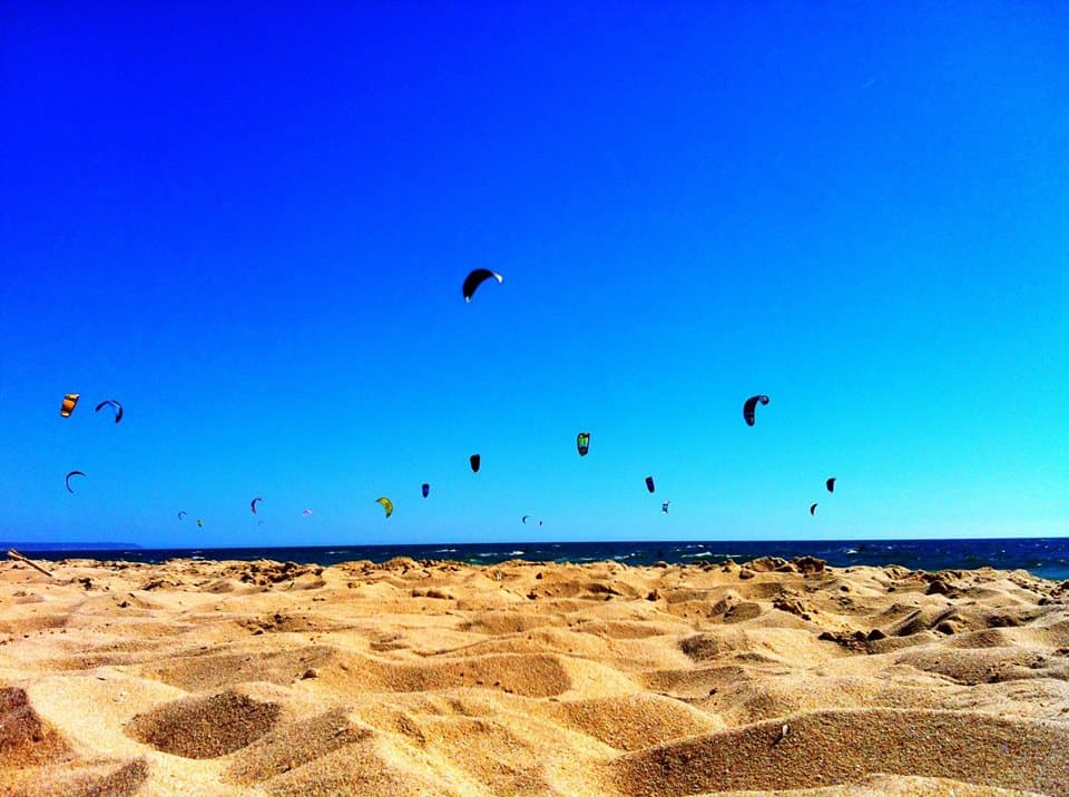 electric blue sky and golden sand of the kitesurfing beach in cascais, portugal. a dozen or so kites fly in the air above the water.