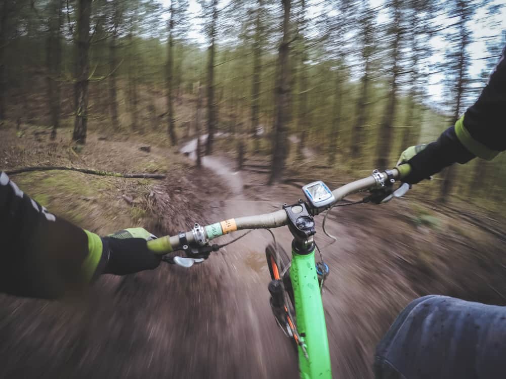 Plan d'ensemble d'une personne faisant de la descente en VTT à travers la forêt sur une piste unique. Le vélo est vert vif et le cycliste porte des gants, des manches noires et un jean.