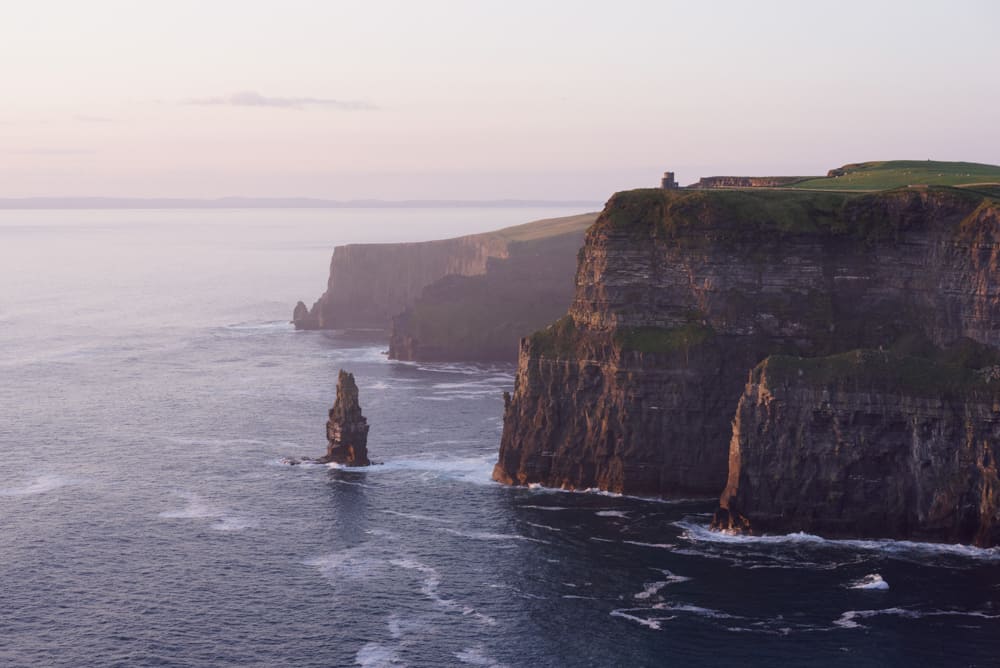 soft pink light at dusk at the cliffs of moher in ireland. The cliffs are covered with green grass and rocky faces on the right of the picture, while the swirling water of the sea covers the left.