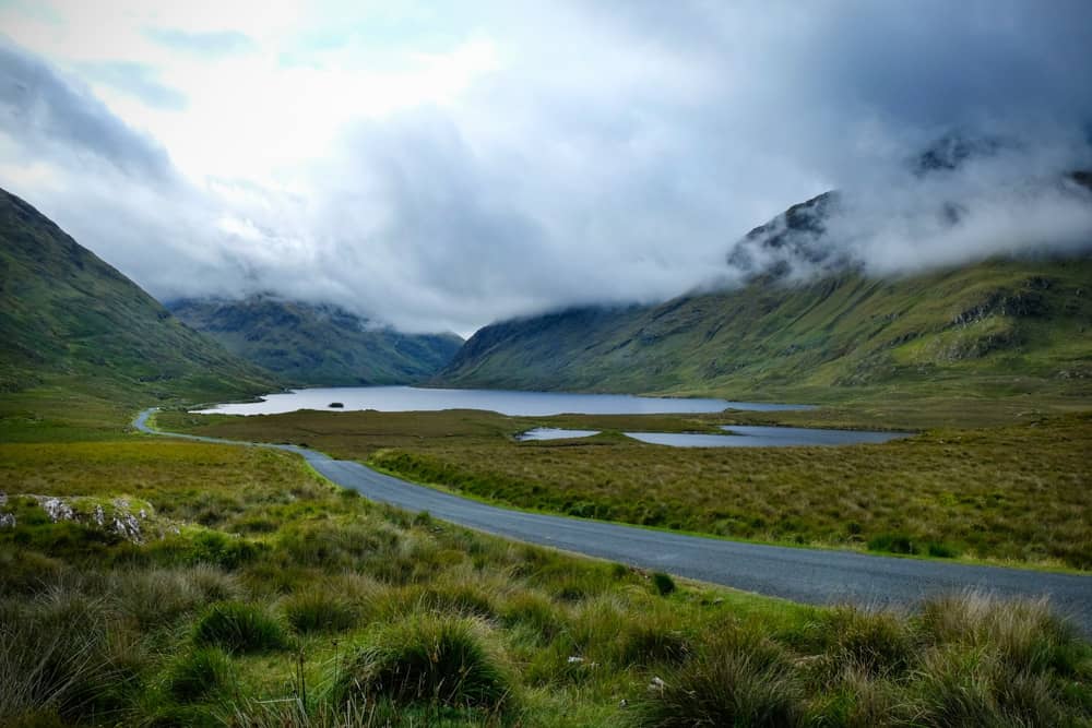 de la brume et des nuages survolent les sommets de montagnes verdoyantes et herbeuses. une route serpente du côté droit au premier plan jusqu'à un lac argenté à l'arrière-plan.