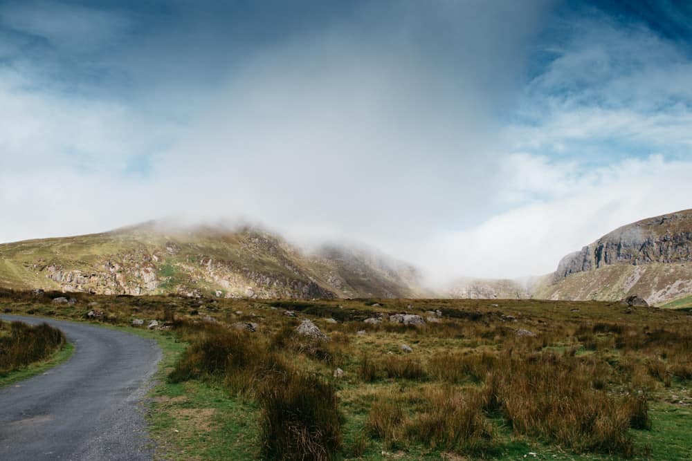 light fluffy clouds hang over the top of some rugged hills in the irish countryside. a small rural road snake across the foreground on the lefthand side.