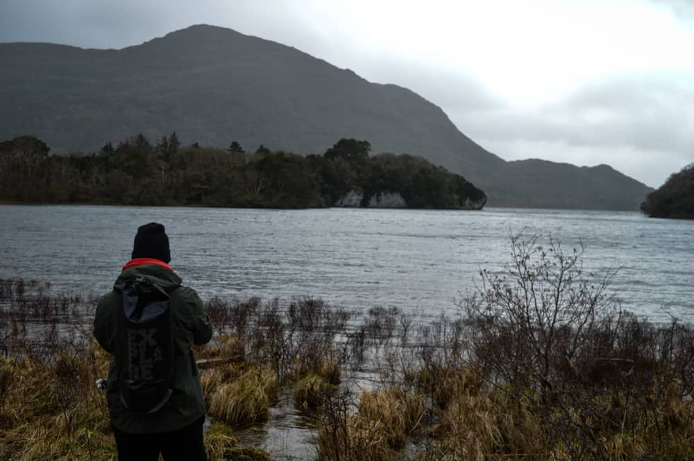 eine Person mit Hut, Regenmantel und Rucksack steht mit dem Rücken zur Kamera und blickt auf einen See und eine Berglandschaft im Killarney National Park an einem grauen, bewölkten Tag