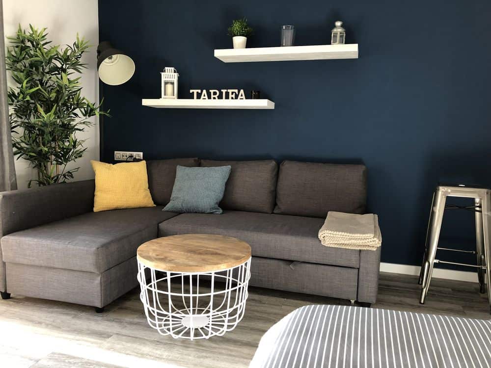 corner of a living room, featuring navy blue walls, a grey couch and coffee table, and white shelves with some candles and a small sign that reads 'tarifa'