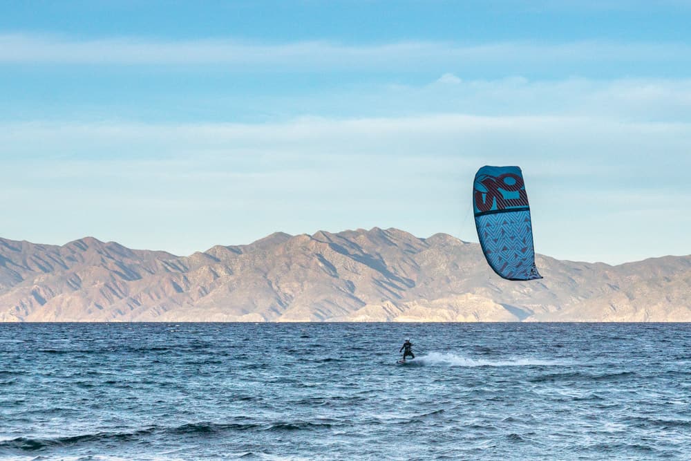 kitesurfer in een wetsuit renners van rechts naar links, vliegend hun blauwe vlieger boven. op de achtergrond zijn er bergen tegen een lichtblauwe hemel