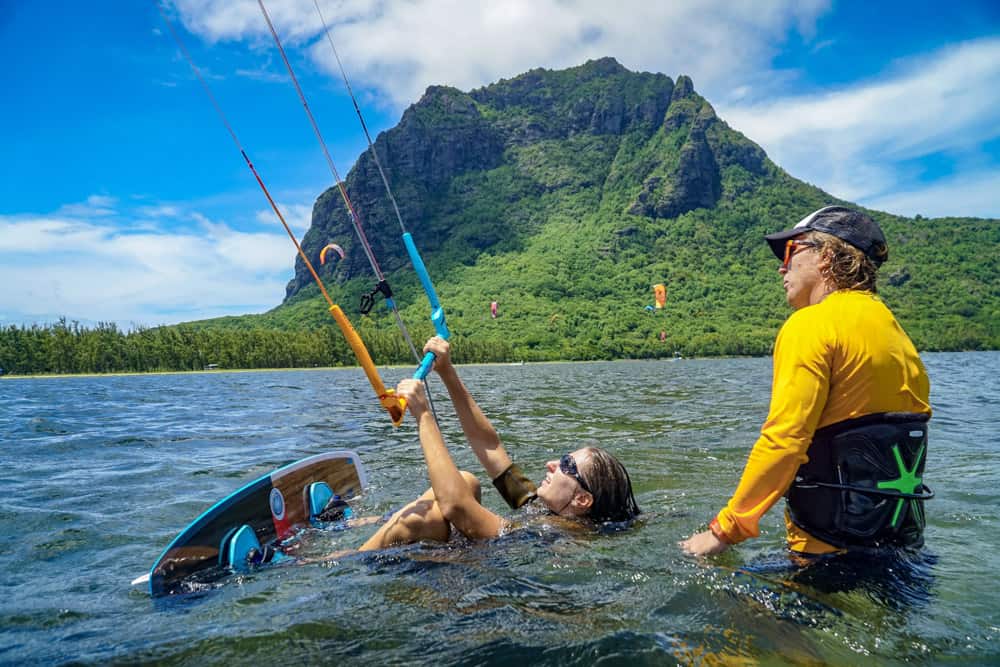 une kitesurfeuse et son instructeur dans l'eau devant une montagne couverte de jungle. l'élève est allongée dans l'eau et se prépare à diriger son aile pour un départ dans l'eau.