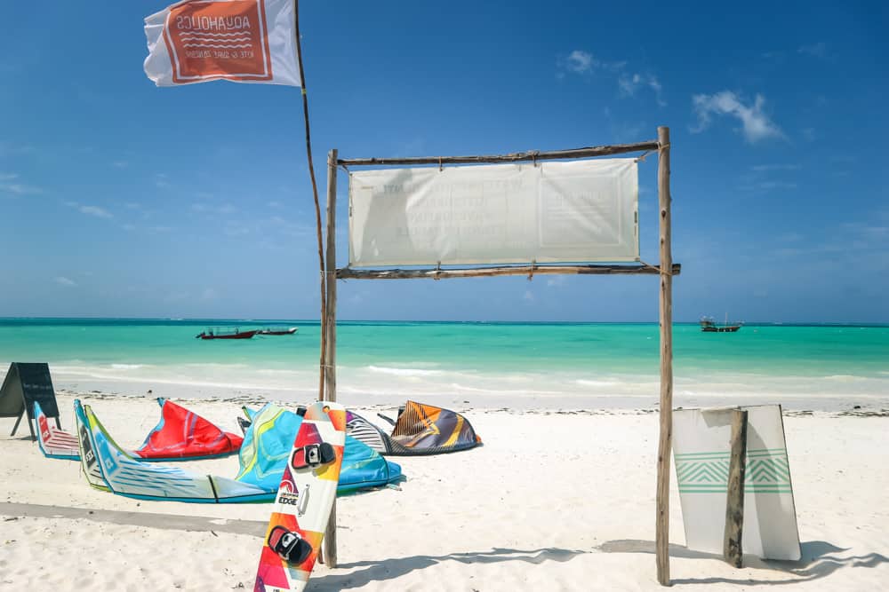 windiger kitesurfing-strand in sansibar, tansania. weißer sand bedeckt den vordergrund, in dem 3 bunte drachen liegen. ein brett und eine flagge lehnen an einem schild, das über azurblaues wasser hinausschaut.