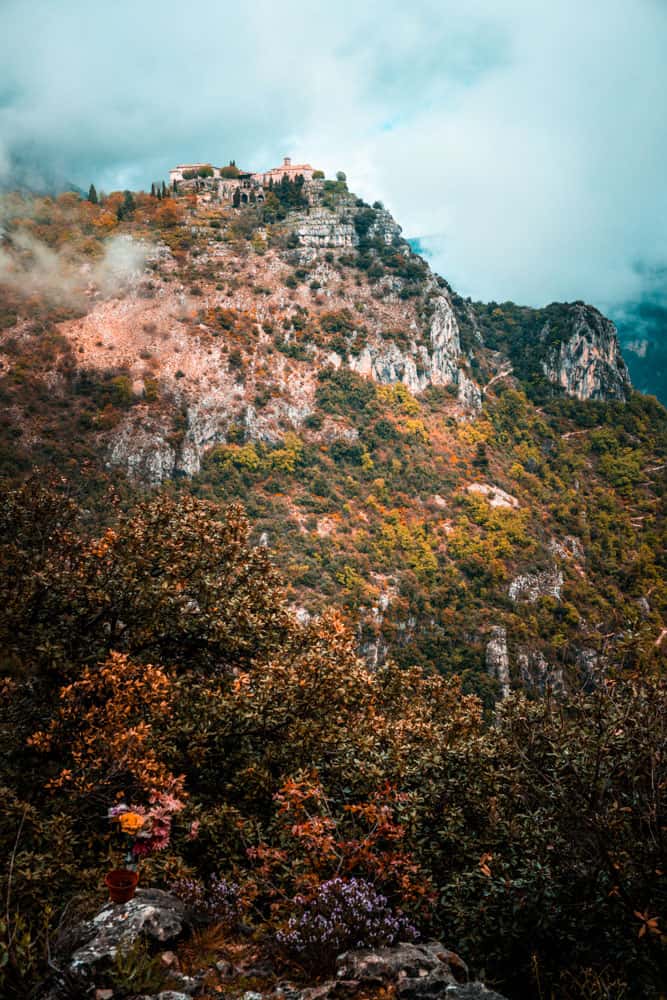 The village of Gourdon perched up on top of a misty cliff. Clouds enshroud some of the village and a portion of the mountain. The cliff face is tinged with orange and lush green foliage. Some wildflowers are growing in the foreground of the picture