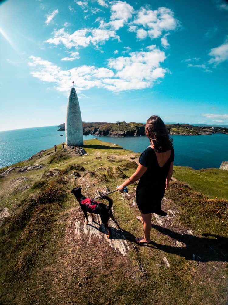 Grace and Blacky the dog standing on a cliff facing the Baltimore Beacon, County Cork. The blue sky is filled with fluffy white clouds and the scene overlooks Sherkin island.