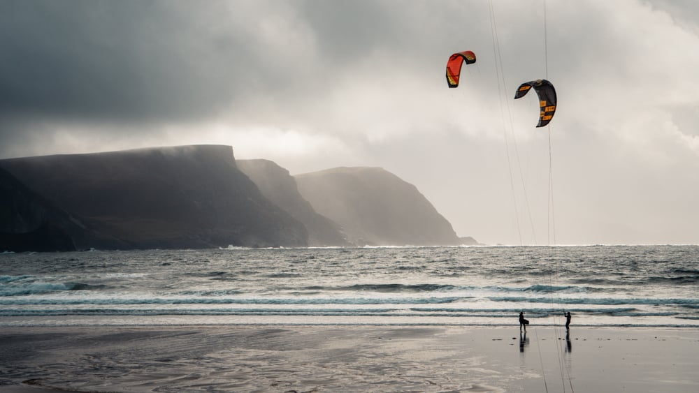 Two kitesurfers standing with their kites in the air on Keel Beach, Achill Island, County Mayo, Ireland. The misty mountains in the back left of the photo are almost totally enshrouded in cloud. It's a grey, wet day.