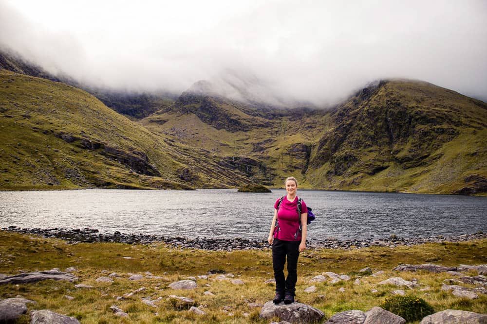 Eine Frau in einem rosa T-Shirt steht vor der nebligen Landschaft des Carrauntoohil, dem höchsten Berg Irlands
