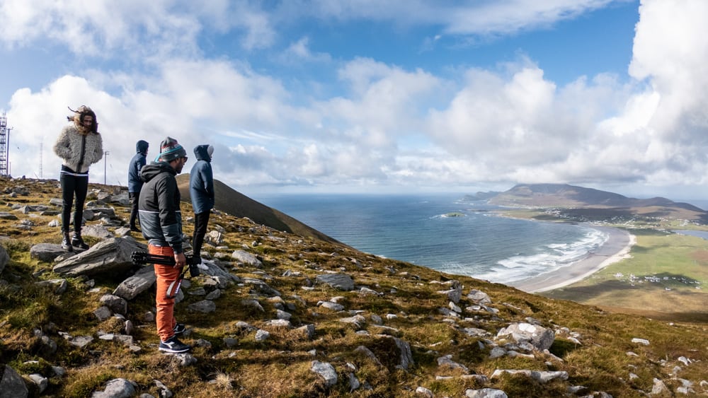 vue du sommet de minaun heights, sur l'île d'achill. quatre personnes sont debout et regardent la vue, qui s'étend des vagues de la plage de keel jusqu'à la péninsule voisine. le ciel est bleu avec des nuages blancs.