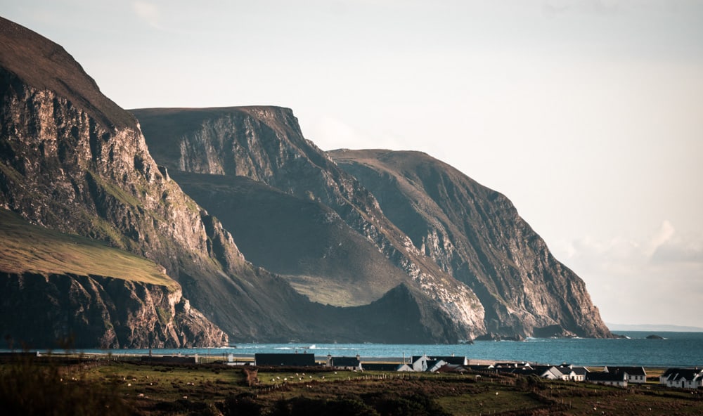 dramatic sea cliffs at keel beach, achill island, county mayo, ireland. The water in the bay is bright blue and the cliffs are lit up by the sun at golden hour. A few houses lie at the foot of the cliffs.