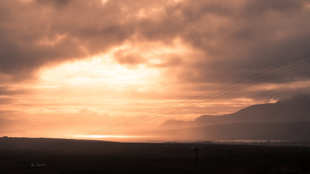 tangerine sunset on achill island, county mayo. the sky is filled wihtmist and clouds, the sun is barely shining through. sea cliffs can just be seen in silhouette.