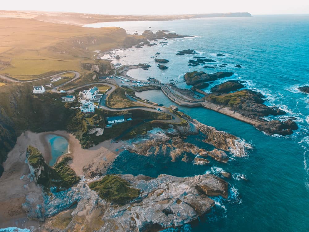 Luftaufnahme von Ballintoy Harbour im Norden von Irland. Das Wasser ist wunderschön blau und ein Lichtschein kriecht von links oben über die Küstenlandschaft. Eine gewundene Straße schlängelt sich durch eine Handvoll Häuser in der Nähe der zerklüfteten Küstenlinie.