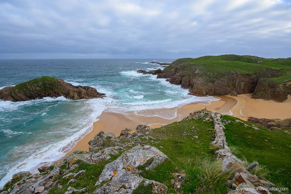 Murder Hole Beach as seen from the hills above. Blue water washes in and meets with the golden sand of a small cove. It's surrounded by grassy green hills. The weather looks stormy.