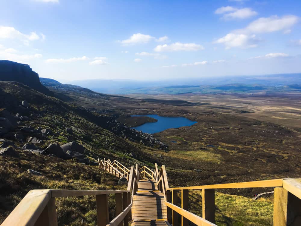 Ireland's Stairway to Heaven, van bovenaf gefotografeerd. De houten trap kruipt omhoog langs de zijkant van een steile berg en kijkt uit over een meer en een uitgestrekt landschap.