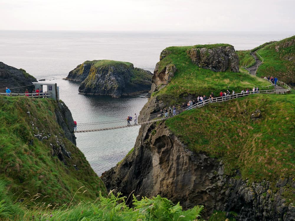 Carrick-a-rede Seilbrücke von einem Aussichtspunkt aus. Viele Menschen überqueren die spindeldürre Brücke, die zwei grüne Grashügel miteinander verbindet, die durch eine Höhle getrennt sind.