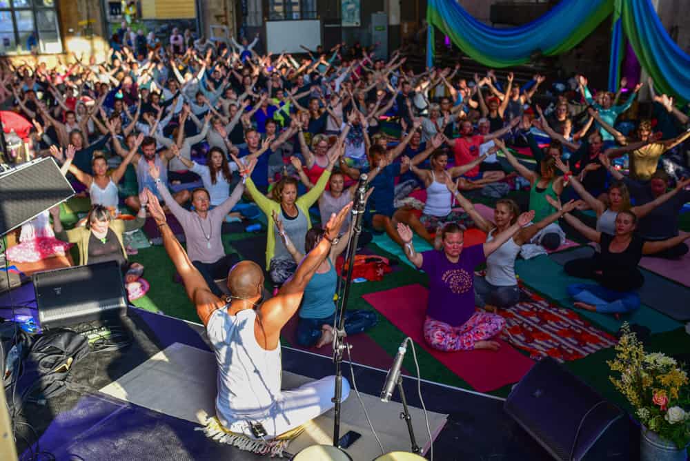 a crowd of people seated on yoga mats doing asanas while led by a teacher on a stage