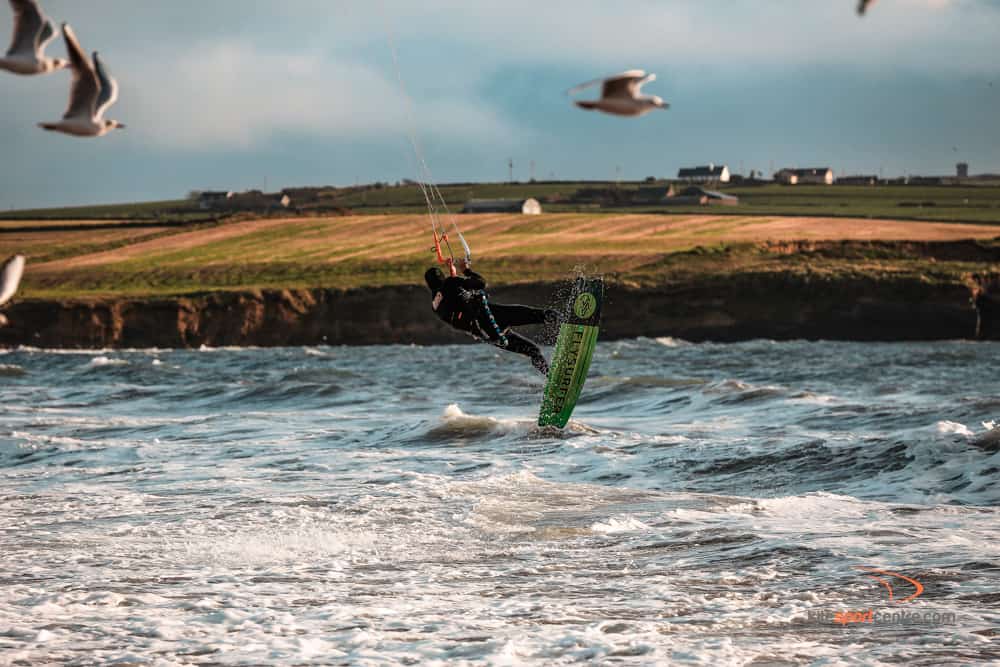 a kitesurfer wearing a black wetsuit jumps over the water. seagulls are flying around him and we can see farmland in the background.