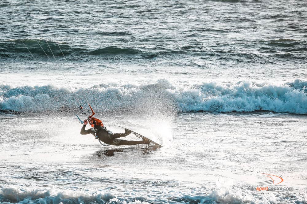 ein schwarz-orange gekleideter Kitesurfer zieht einen Darkslide in der flachen Zone zwischen den Wellen