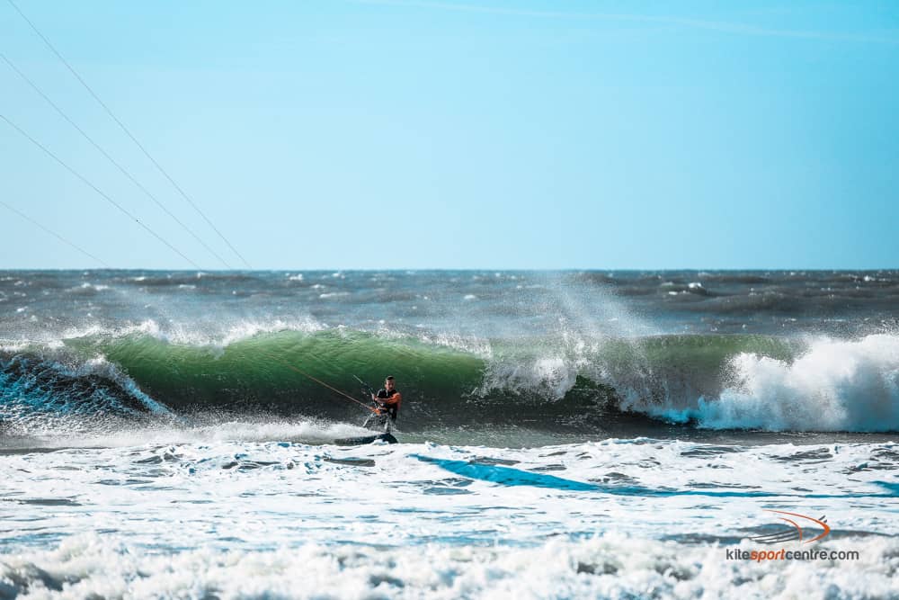 un kitesurfeur portant une combinaison noire et orange traverse les vagues par une journée agitée en irlande. une vague se brise juste derrière lui.