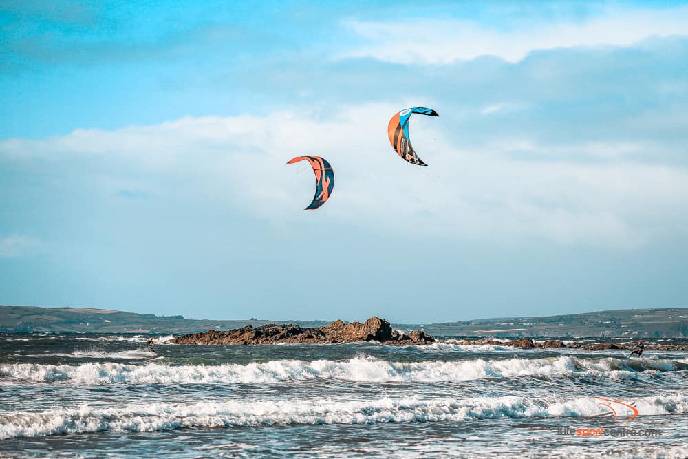 two kitesurfers riding towards each other in the choppy irish waters. there are craggy rocks sticking out of the water and the coastline is just visible behind.